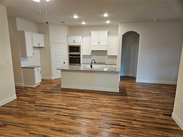 kitchen featuring stainless steel appliances, dark wood finished floors, a center island with sink, arched walkways, and white cabinets