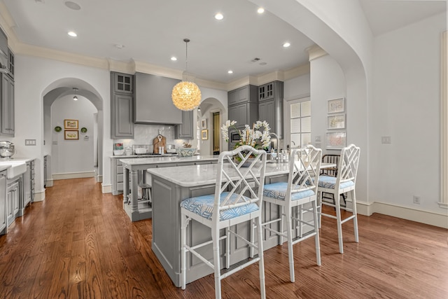 kitchen featuring gray cabinets, a large island with sink, glass insert cabinets, wall chimney range hood, and arched walkways