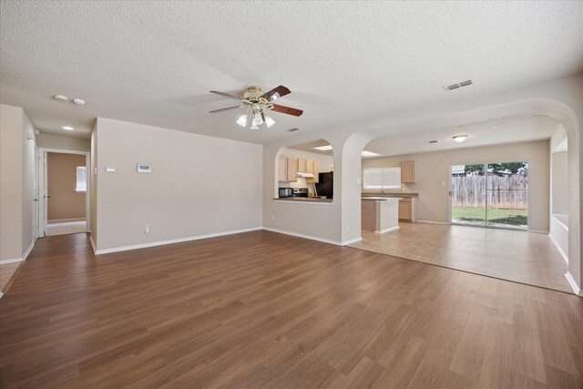 unfurnished living room featuring arched walkways, a textured ceiling, a ceiling fan, wood finished floors, and baseboards