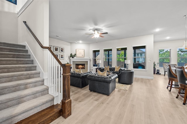 living area featuring light wood-type flooring, a ceiling fan, a warm lit fireplace, recessed lighting, and stairs
