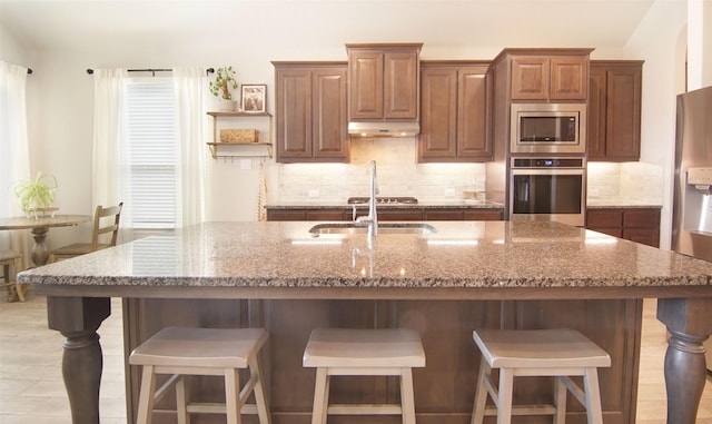 kitchen with stainless steel appliances, under cabinet range hood, decorative backsplash, a sink, and light stone counters