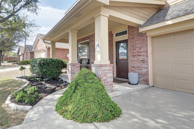 view of exterior entry with an attached garage, driveway, brick siding, a porch, and a shingled roof