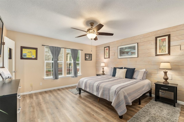 bedroom with a textured ceiling, wooden walls, ceiling fan, light wood-style flooring, and baseboards