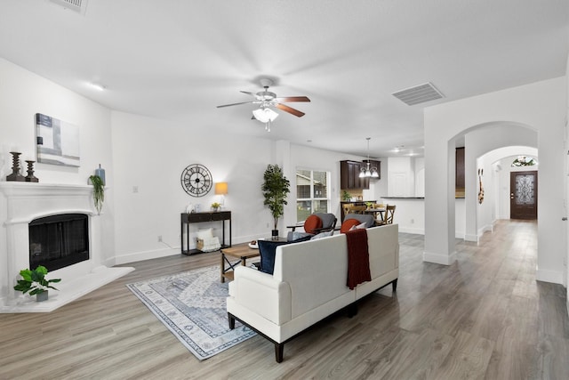 living area featuring visible vents, arched walkways, a fireplace with raised hearth, ceiling fan with notable chandelier, and light wood-style floors