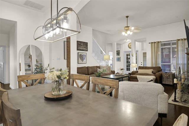 dining space with arched walkways, dark wood finished floors, visible vents, ceiling fan with notable chandelier, and stairs