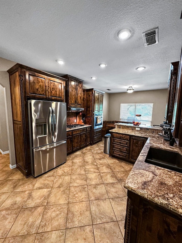kitchen with stainless steel appliances, dark brown cabinets, a textured ceiling, recessed lighting, and dark stone counters