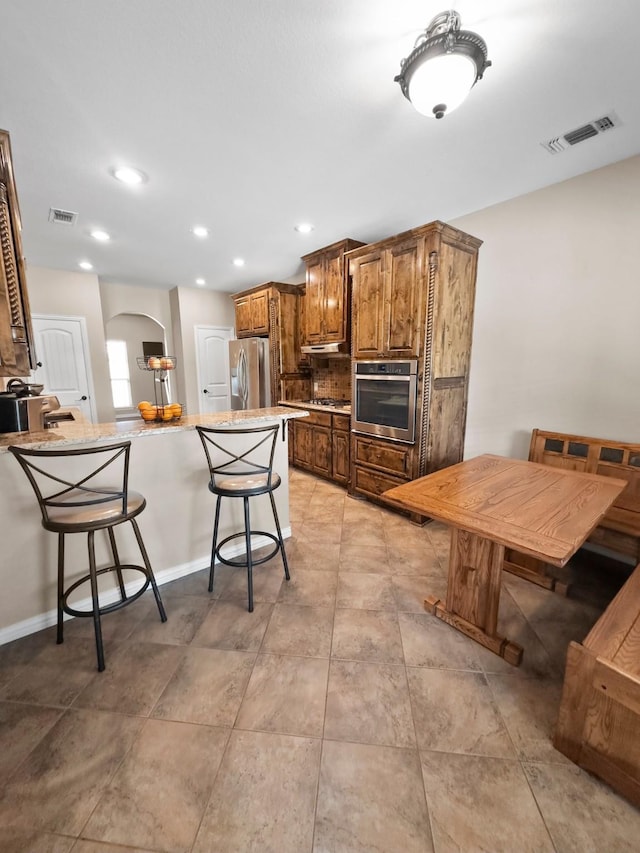 kitchen featuring appliances with stainless steel finishes, a kitchen bar, arched walkways, brown cabinetry, and recessed lighting