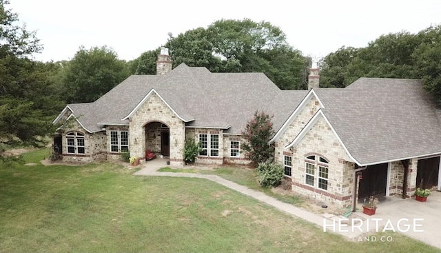 french country home featuring a front yard, stone siding, roof with shingles, a chimney, and an attached garage