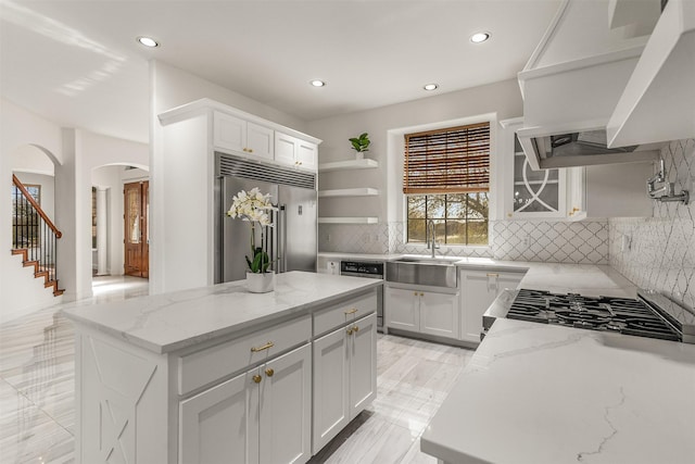 kitchen with open shelves, light stone countertops, a sink, arched walkways, and built in fridge
