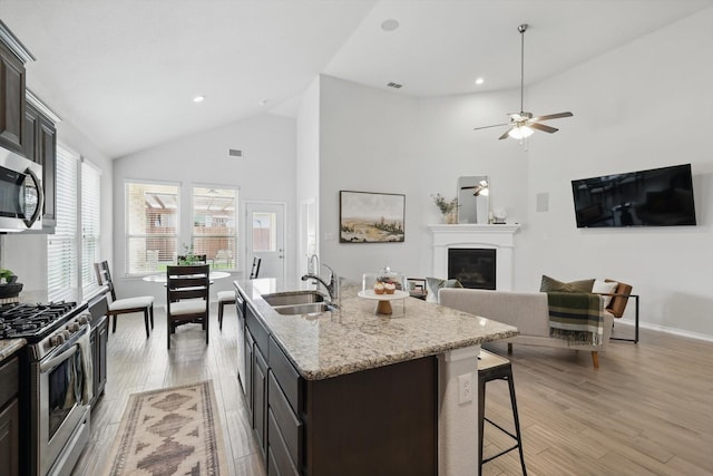 kitchen with a sink, a ceiling fan, stainless steel appliances, high vaulted ceiling, and a glass covered fireplace