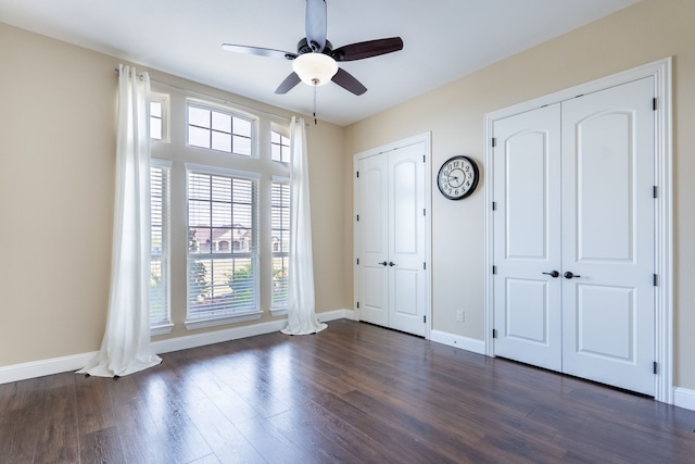 unfurnished bedroom featuring dark wood-type flooring, two closets, baseboards, and ceiling fan