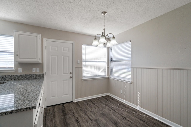 unfurnished dining area with wainscoting, a textured ceiling, a notable chandelier, and dark wood-style flooring