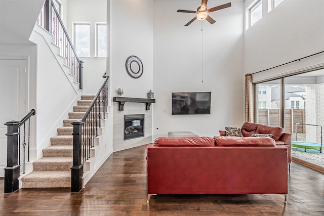 living area featuring ceiling fan, wood finished floors, a glass covered fireplace, stairway, and a high ceiling