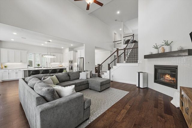 living room featuring stairs, dark wood-style flooring, a high ceiling, ceiling fan, and a glass covered fireplace
