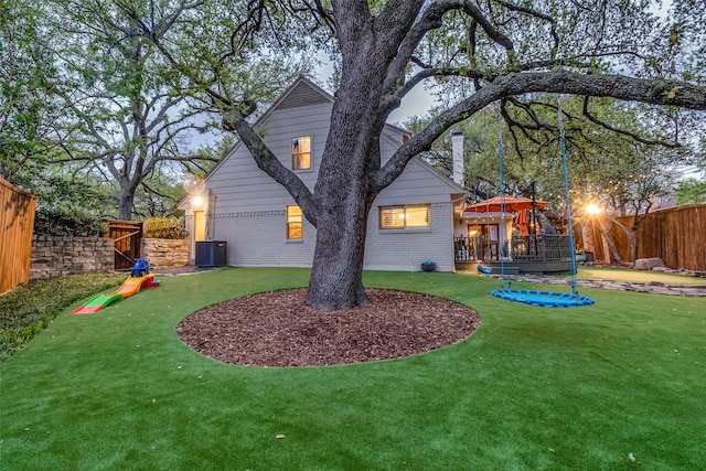 rear view of property featuring a playground, central AC unit, a fenced backyard, a lawn, and brick siding