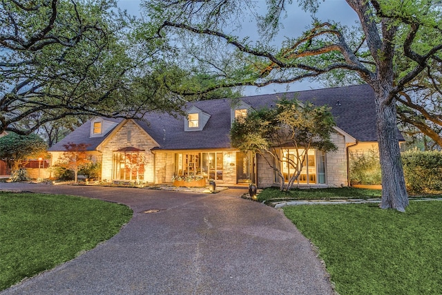 new england style home with roof with shingles, stone siding, a front yard, and curved driveway