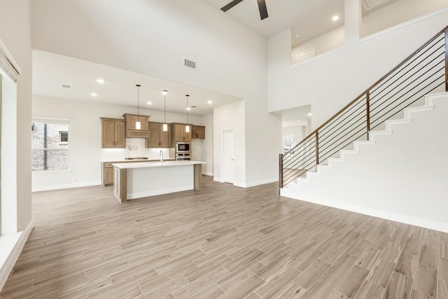 unfurnished living room featuring stairs, light wood-type flooring, ceiling fan, baseboards, and a towering ceiling