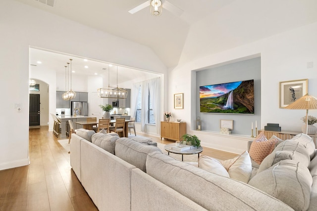 living area featuring light wood finished floors, vaulted ceiling, baseboards, arched walkways, and ceiling fan with notable chandelier