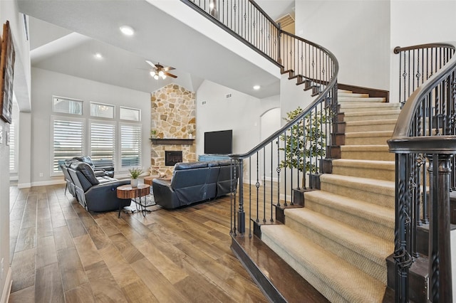 living room featuring stairs, ceiling fan, wood finished floors, a stone fireplace, and a high ceiling