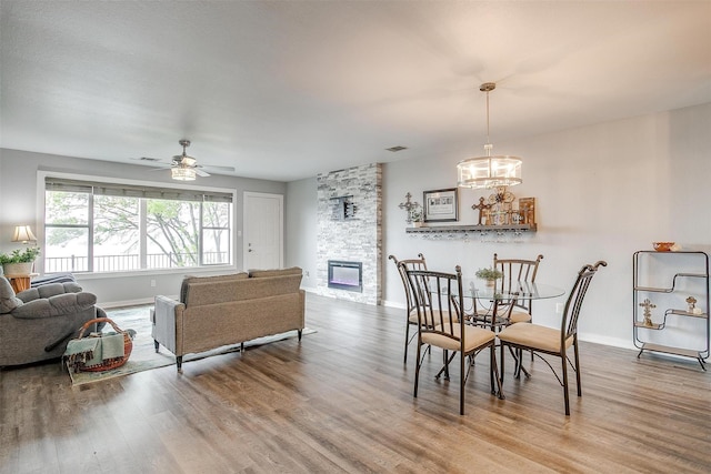 dining room featuring ceiling fan with notable chandelier, a stone fireplace, wood finished floors, and baseboards