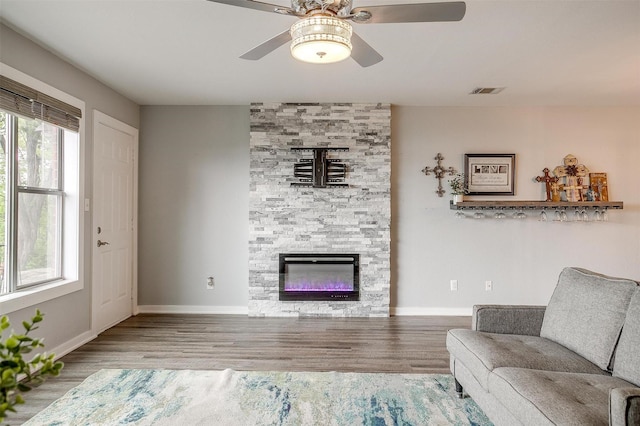 living area featuring wood finished floors, visible vents, a stone fireplace, baseboards, and ceiling fan