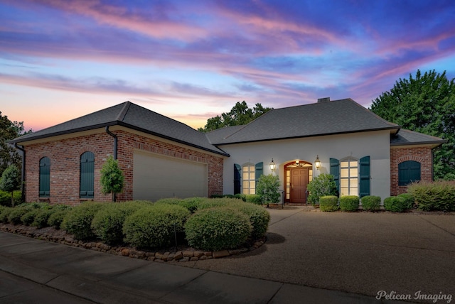 french provincial home featuring driveway, stucco siding, an attached garage, brick siding, and a shingled roof