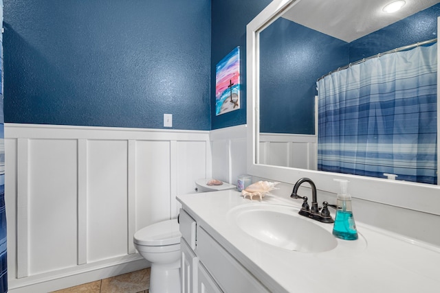 full bath featuring toilet, vanity, a shower with curtain, a textured wall, and wainscoting