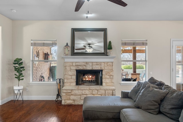 living room with a stone fireplace, dark wood-style floors, ceiling fan, and baseboards