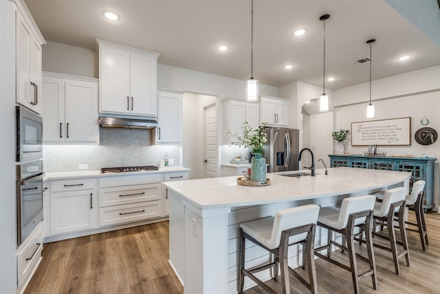 kitchen featuring decorative backsplash, white cabinets, a kitchen island with sink, appliances with stainless steel finishes, and under cabinet range hood
