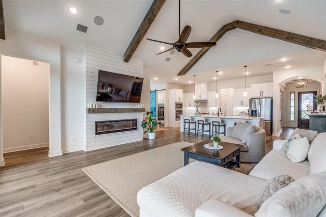living area featuring arched walkways, beamed ceiling, light wood finished floors, visible vents, and a ceiling fan