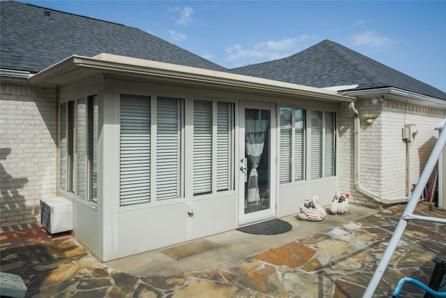view of exterior entry featuring a patio, brick siding, and roof with shingles