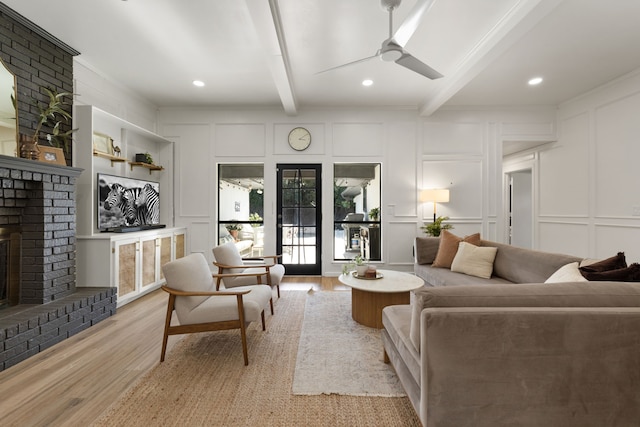 living room featuring a brick fireplace, light wood-style flooring, beamed ceiling, ceiling fan, and a decorative wall