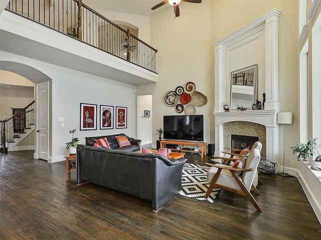 living area with dark wood-style floors, a tiled fireplace, a towering ceiling, and a ceiling fan