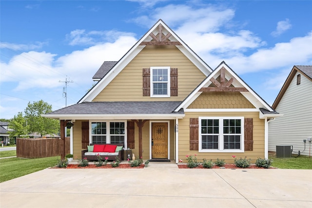 view of front facade with fence, a front lawn, roof with shingles, covered porch, and cooling unit