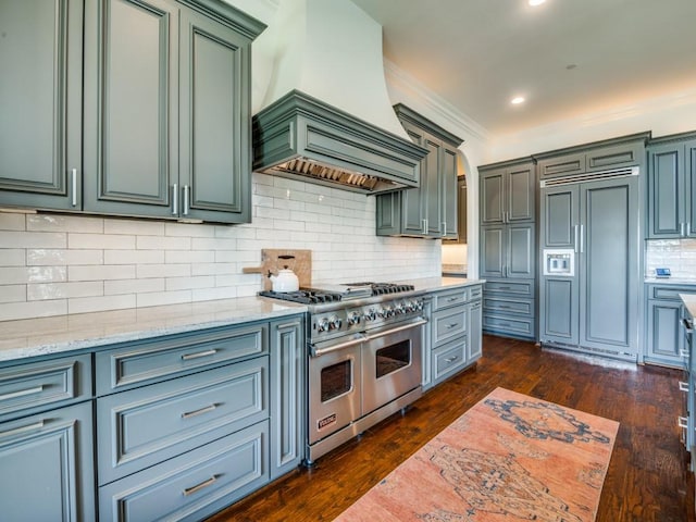 kitchen featuring backsplash, high quality appliances, light stone countertops, premium range hood, and dark wood-type flooring