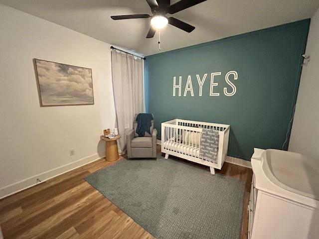 bedroom featuring a ceiling fan, baseboards, a nursery area, and wood finished floors