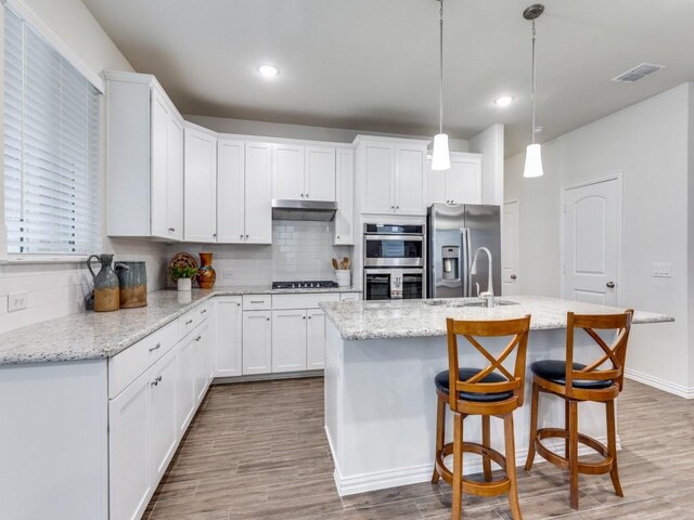 kitchen with a kitchen island with sink, appliances with stainless steel finishes, tasteful backsplash, white cabinets, and under cabinet range hood