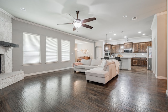 living area featuring visible vents, ornamental molding, a ceiling fan, a stone fireplace, and dark wood finished floors
