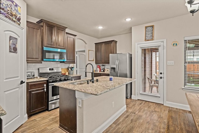 kitchen featuring light wood-style floors, dark brown cabinetry, a kitchen island with sink, a sink, and stainless steel appliances