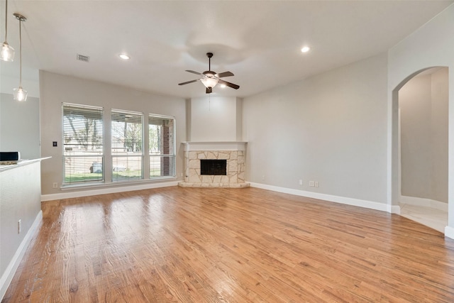 unfurnished living room featuring baseboards, a stone fireplace, light wood-style floors, ceiling fan, and arched walkways