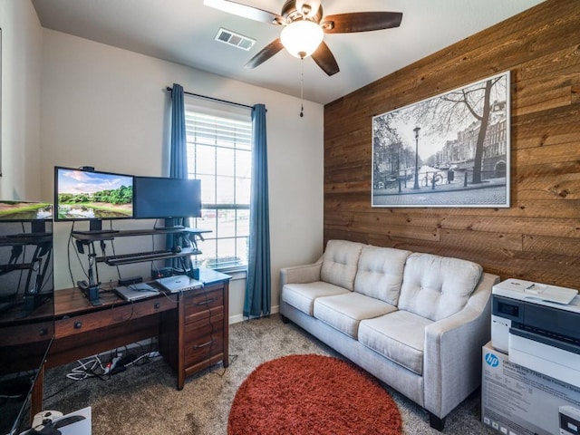 carpeted office with wood walls, visible vents, a ceiling fan, an accent wall, and baseboards