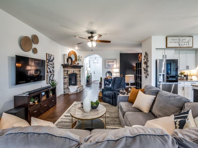 living area featuring baseboards, a stone fireplace, ceiling fan, and dark wood-style flooring