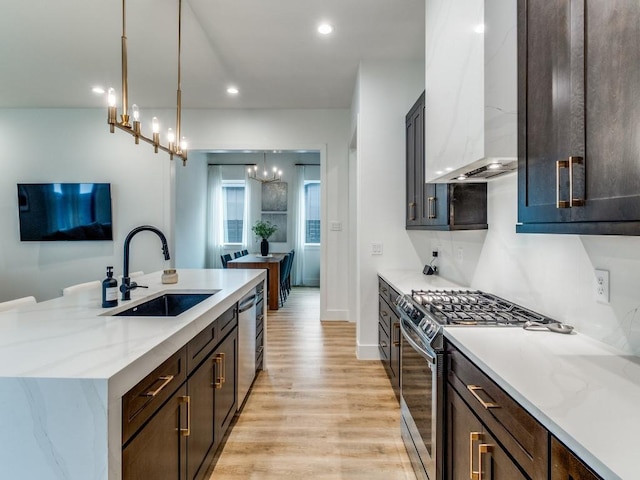 kitchen with an inviting chandelier, stainless steel appliances, a sink, light wood-style floors, and wall chimney range hood
