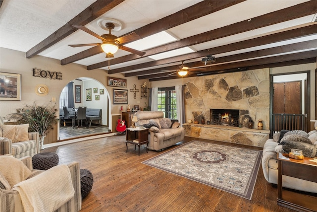 living room with hardwood / wood-style floors, arched walkways, beam ceiling, a ceiling fan, and a stone fireplace