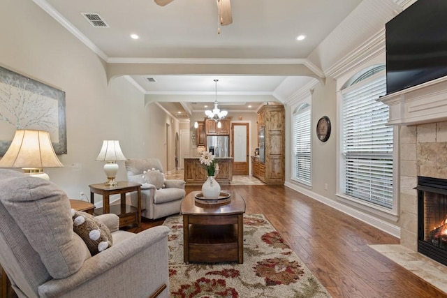 living area featuring a tiled fireplace, baseboards, visible vents, crown molding, and dark wood-type flooring
