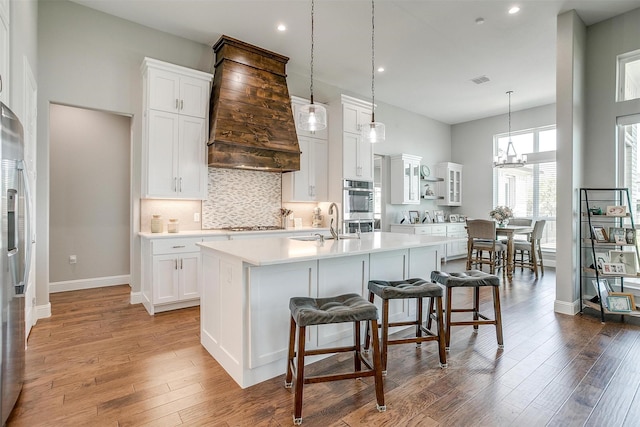 kitchen with custom exhaust hood, light countertops, an island with sink, white cabinetry, and stainless steel appliances