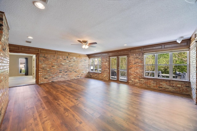 unfurnished living room with ceiling fan, a textured ceiling, visible vents, wood finished floors, and brick wall
