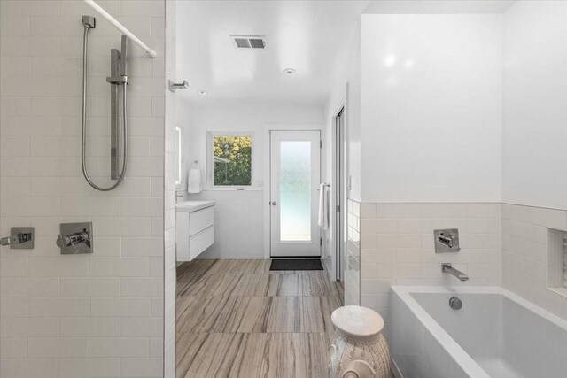 full bathroom featuring a tile shower, a bath, vanity, visible vents, and tile walls