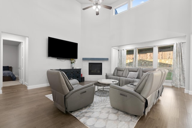 living room with ceiling fan, a high ceiling, a glass covered fireplace, baseboards, and wood finished floors