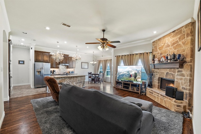 living area with visible vents, ornamental molding, a stone fireplace, dark wood finished floors, and ceiling fan with notable chandelier